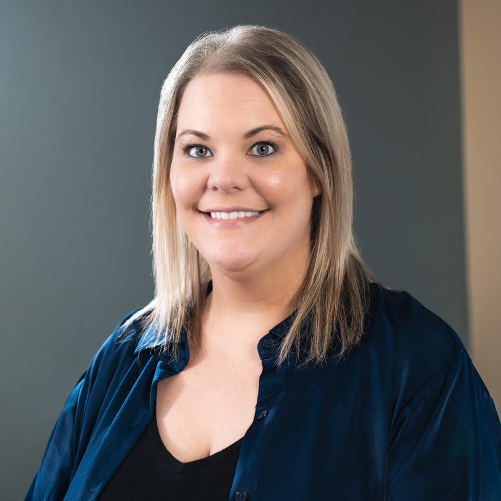 A headshot of Miranda Elliot—a blonde woman wearing a navy blue button up shirt over a black undershirt.