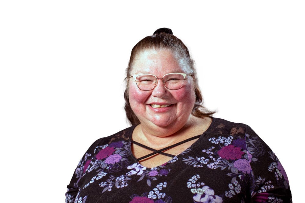 A headshot of Kay Hook—An older white woman with short brown hair, glasses, and a black and pink floral shirt.