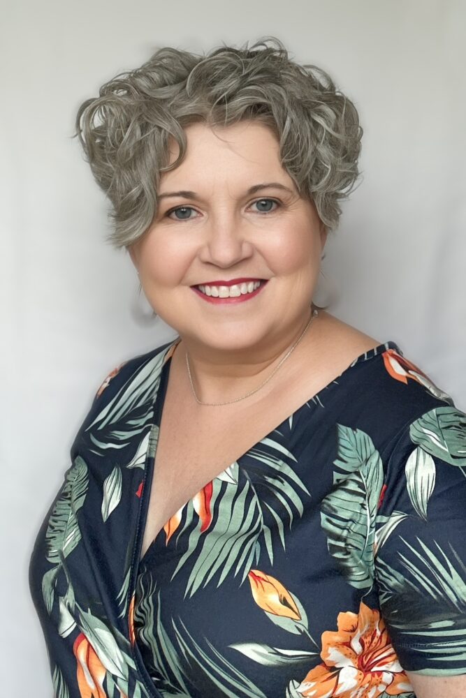 A headshot of Paula Mercer—a woman with short silver curly hair wearing a tropical print dress.