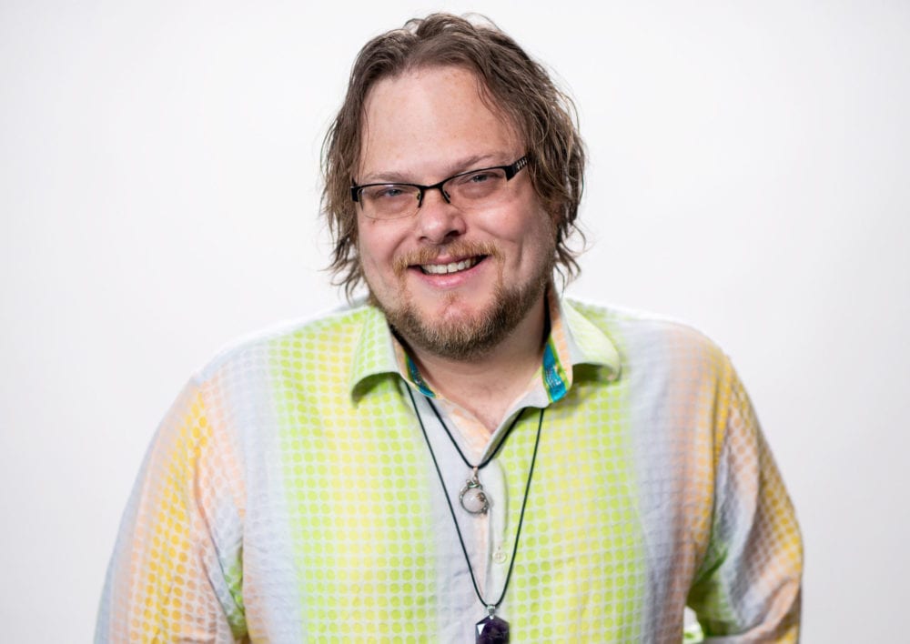 Headshot of Shane Baskins—a white man with longer brown hair, wearing a pastel multicolored polkadot shirt.
