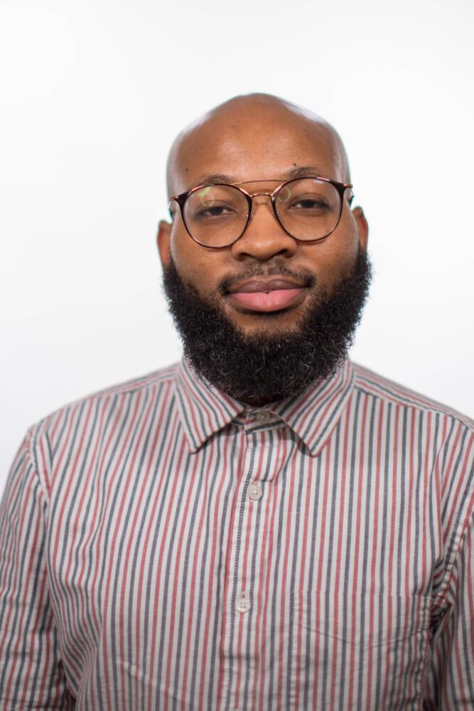 A headshot of Derrick White—a younger black man with a thick beard. He is wearing a red, white, and blue pin striped shirt.
