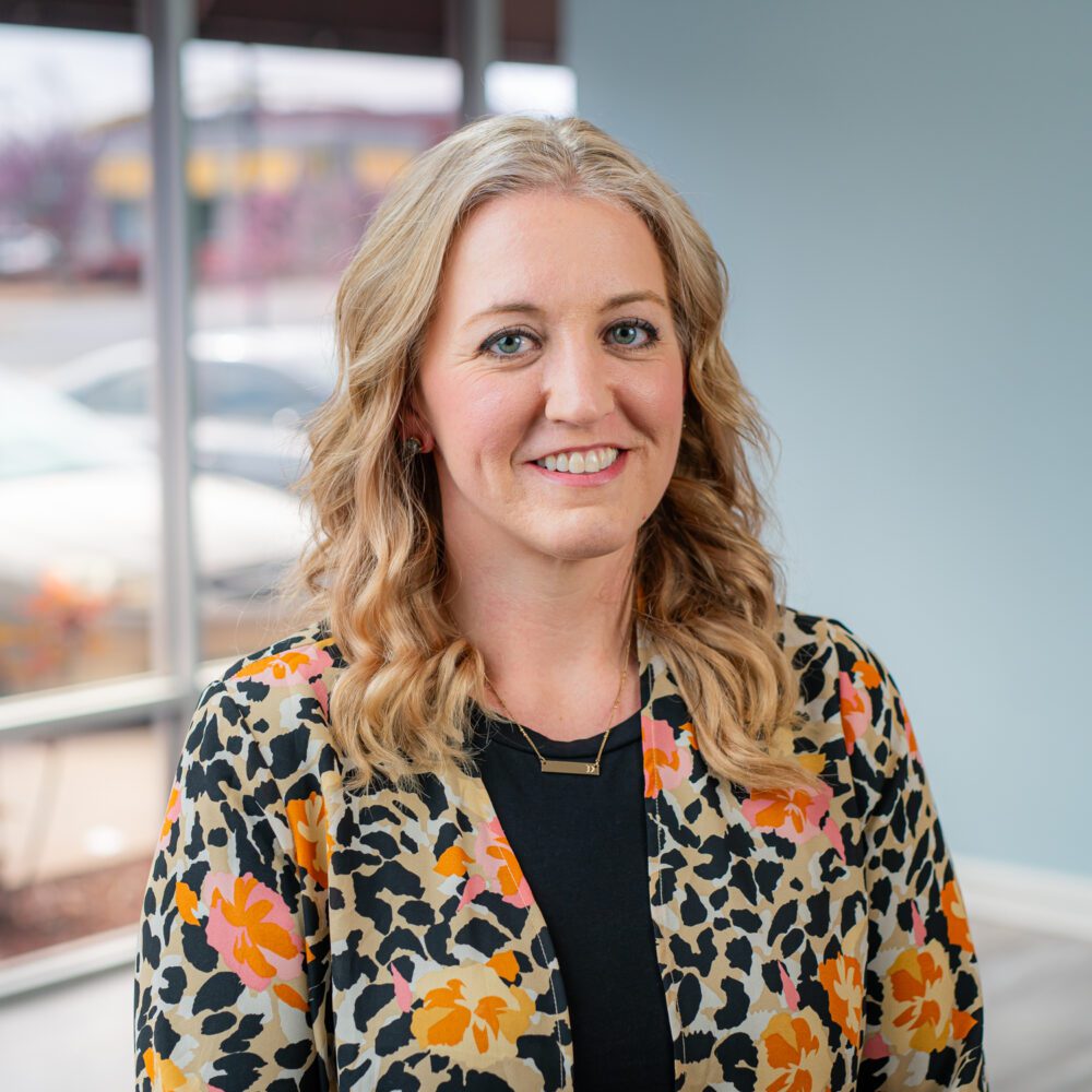 A headshot of Dr. Danielle Bell—a blonde woman wearing a black shirt with a floral print shirt over it.