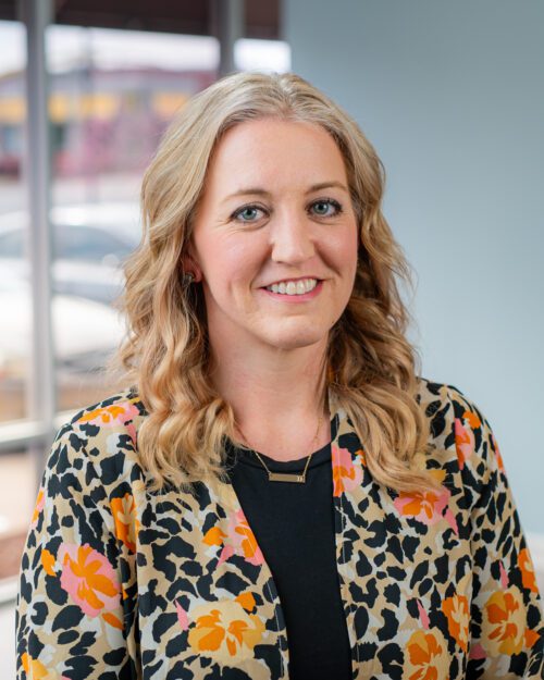 A headshot of Dr. Danielle Bell—a blonde woman wearing a black shirt with a floral print shirt over it.