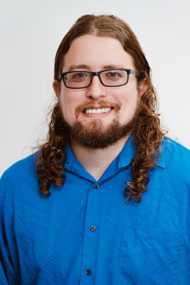 A headshot of Andrew Faught—a younger man with long curly brown hair, wearing a blue button-up shirt.
