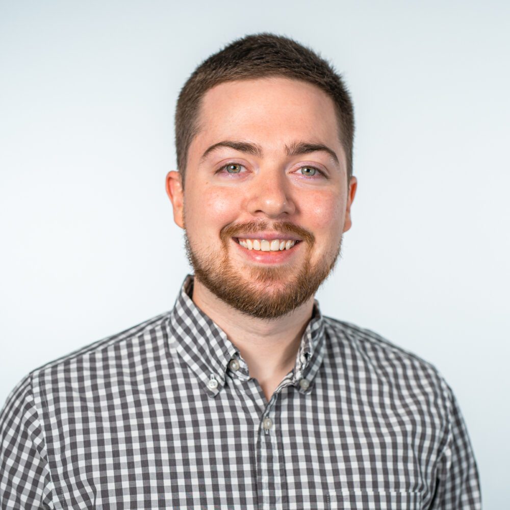 A headshot of Jehu–a younger white man with short brown hair and a beard, wearing a brown plaid button-up shirt.