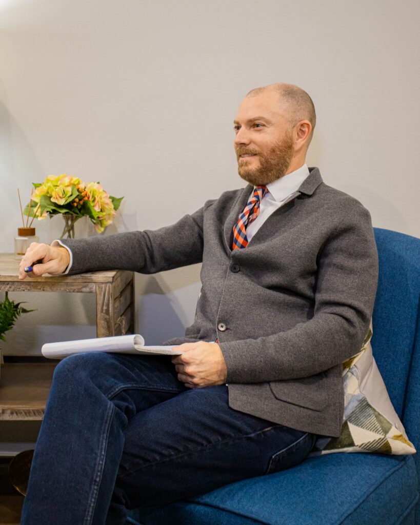 A therapist wearing jeans, a tie, and a gray buttoned jacket, sitting in a blue chair and holding a notepad and blue pen.